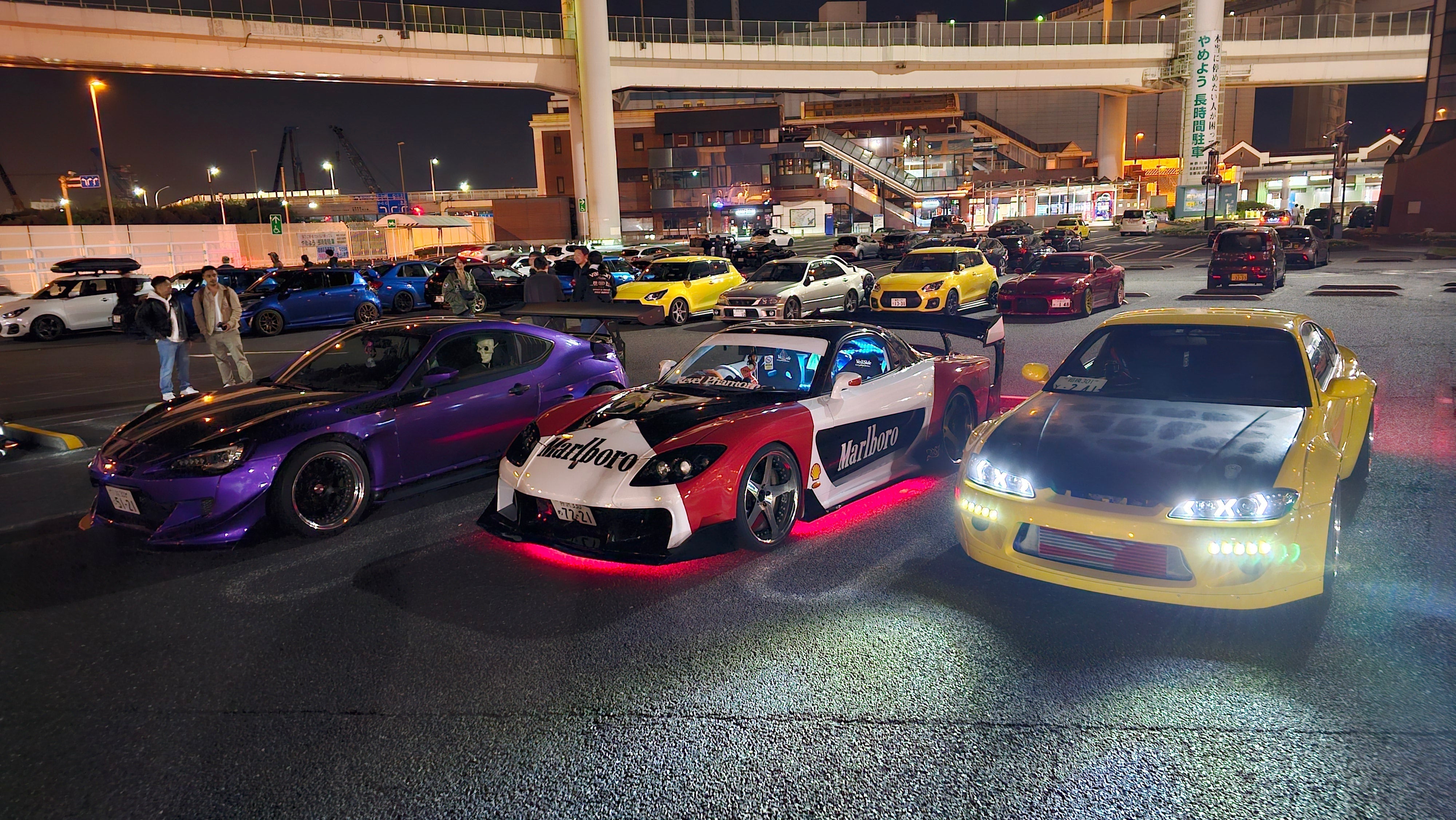A widebody Toyota GT86 in purple next to a Veilside Fortune RX7 in red and white and a yellow Rocket Bunny S15 Silvia sitting at Daikoku Parking Area in Japan at night.